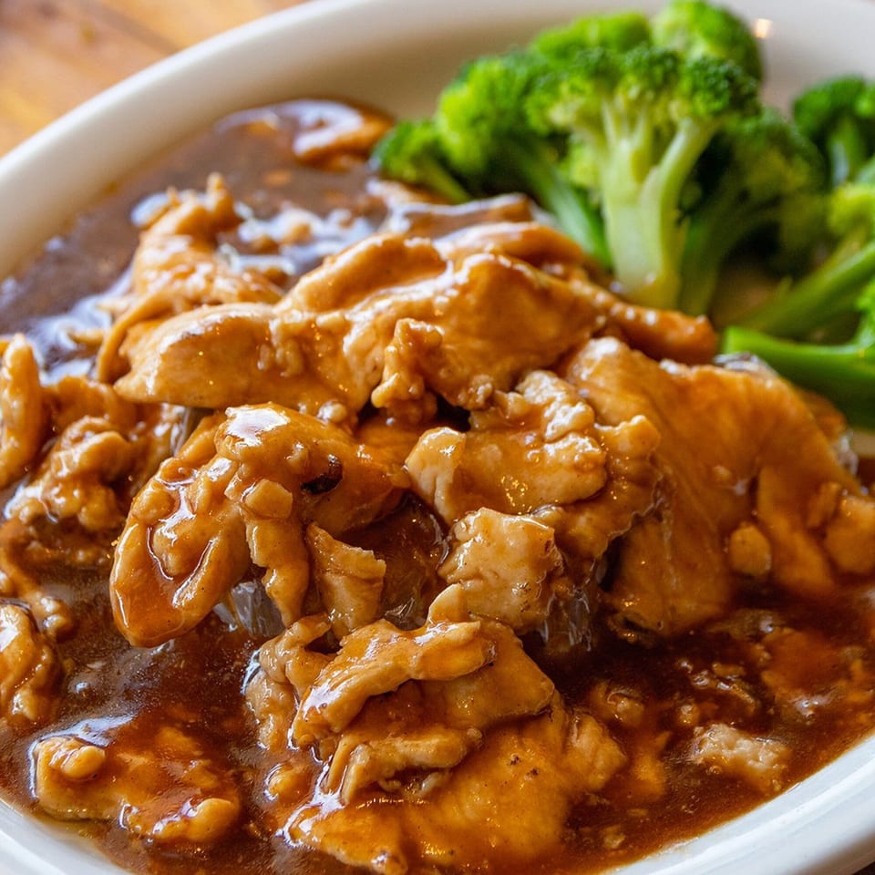 Peppers Thai chicken with brown sauce and a side of steamed broccoli in a white bowl.